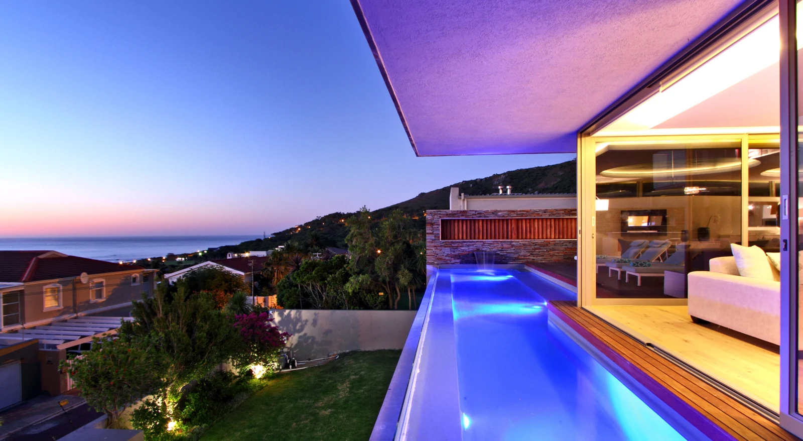 Lit lap pool and terrace of a private villa at dusk, with a sea view and the mountainside beyond