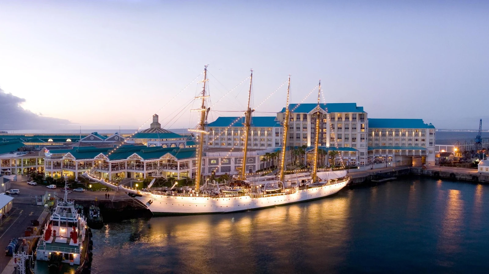 Tall ship moored in the V&A Waterfront harbour at dusk, with the lit harbour buildings behind