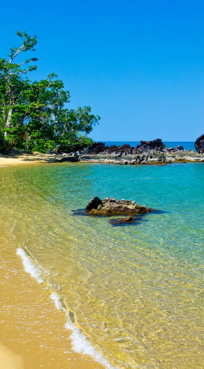 Clear shallow water lapping a sandy shore below forested rocks on the Madagascar coast