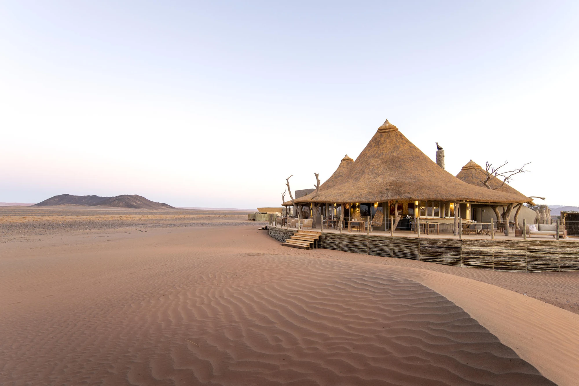Thatched desert lodge on rippled red sand at dawn, with a low mountain on the horizon