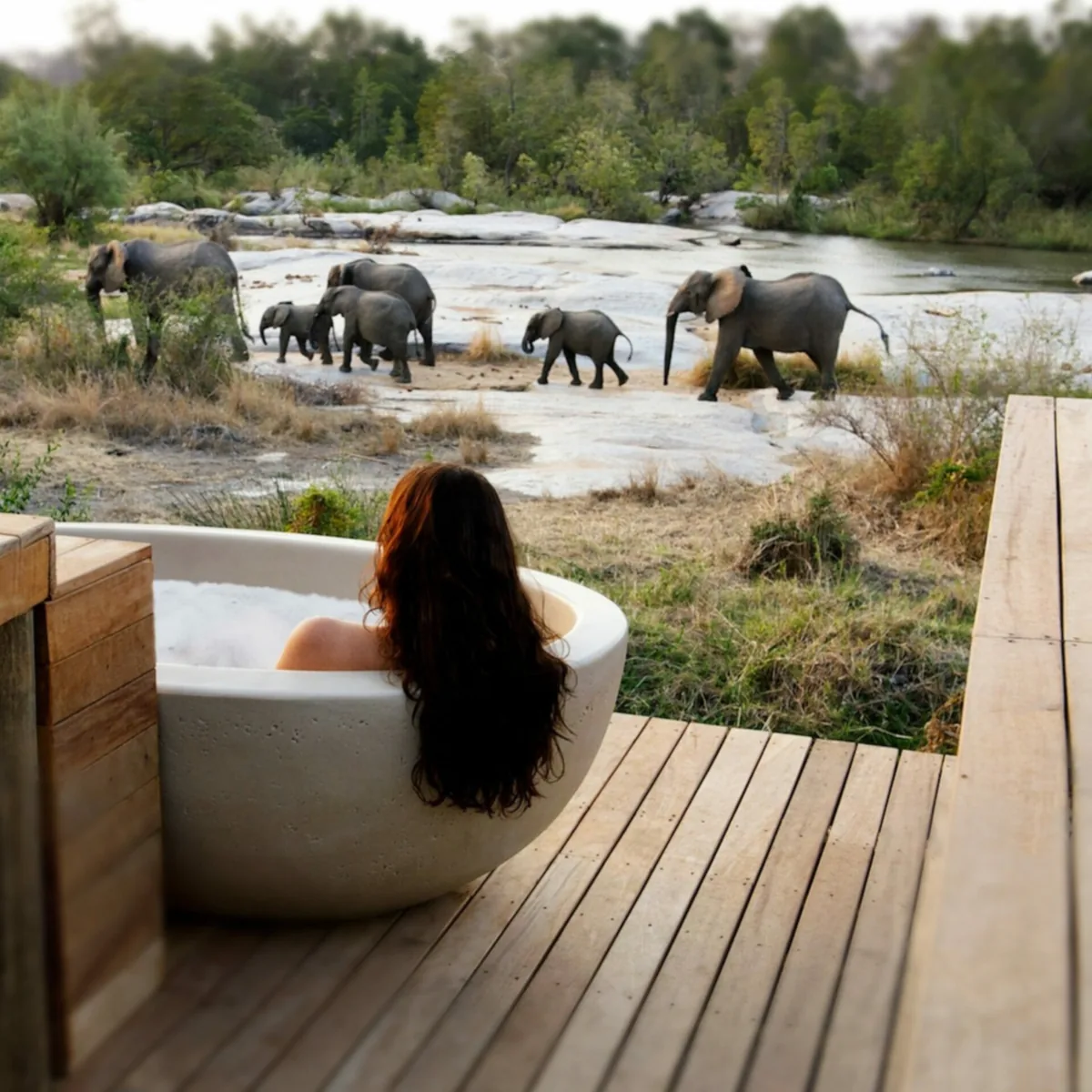 Woman relaxing in an outdoor bathtub on a lodge deck while elephants cross a river