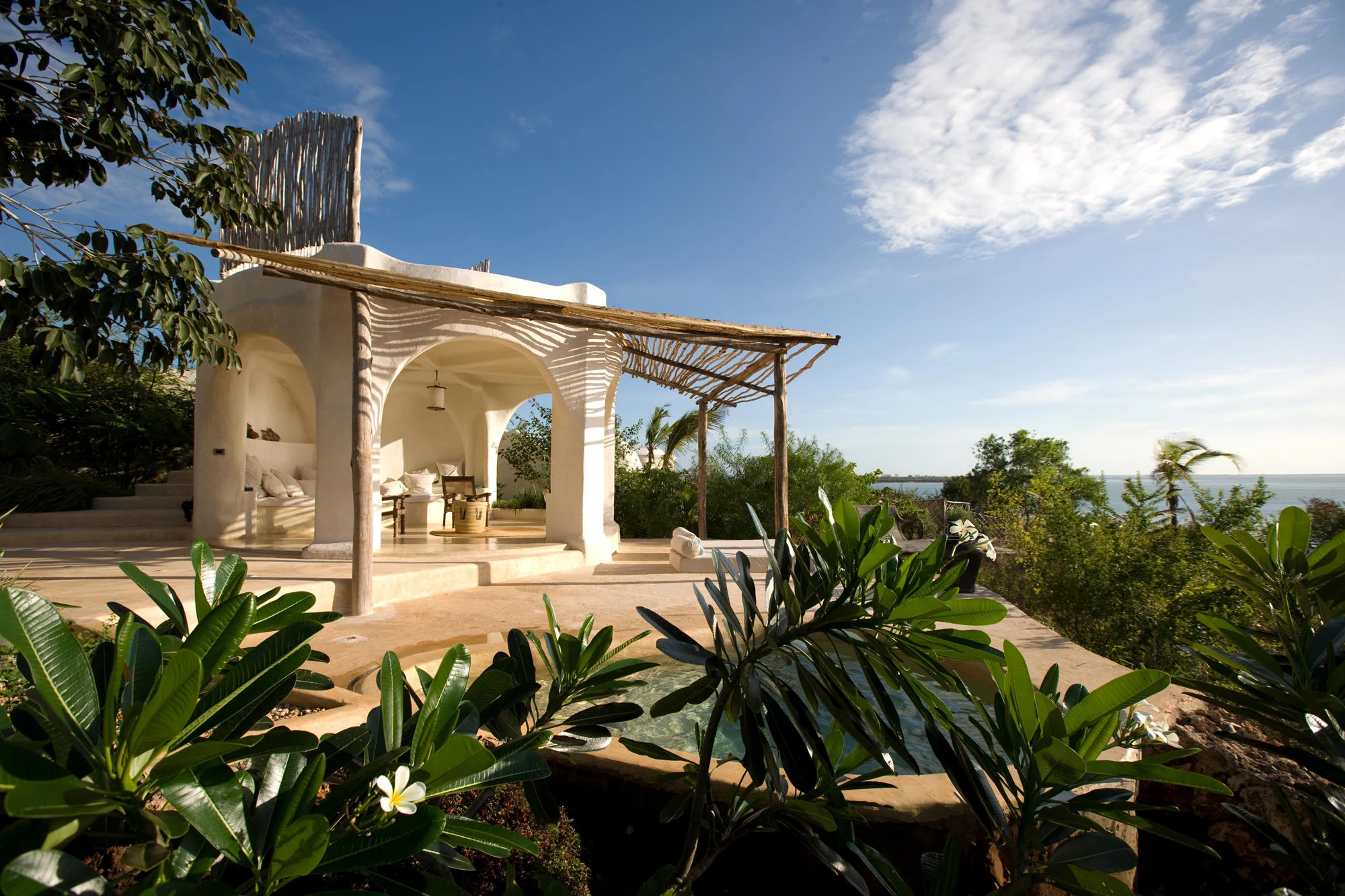 Open-air villa lounge with white arches, a plunge pool and tropical plants, with the Indian Ocean beyond
